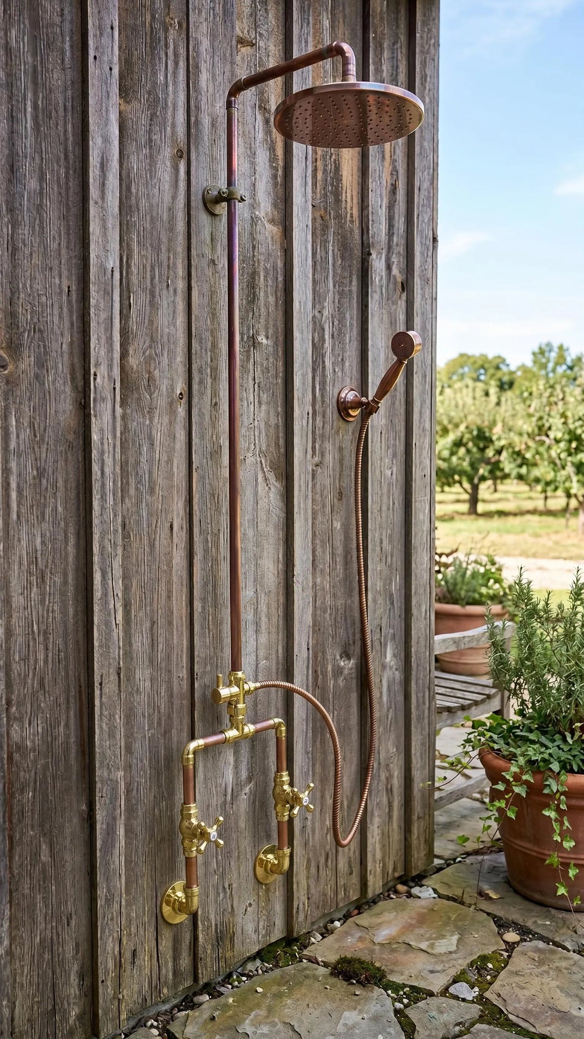 Rustic outdoor shower system with a round overhead shower head, handheld shower hose, and brass valves mounted on a weathered wooden wall beside a stone patio.