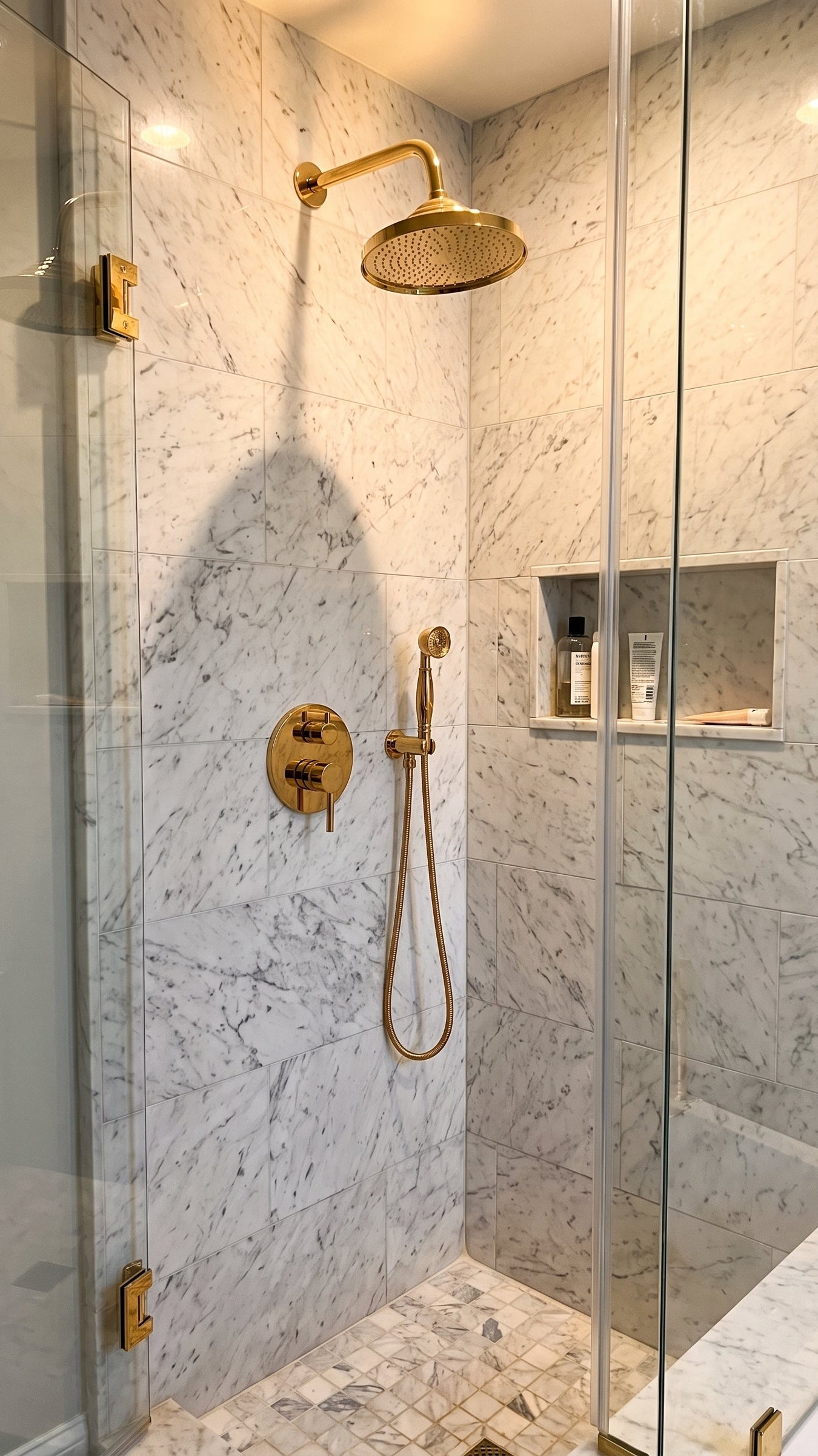 A modern walk-in shower enclosure with a large round gold rain shower head, handheld gold shower wand, and gold control valves. The walls are covered in light gray and white marble tiles, and a recessed shelf holds toiletries.