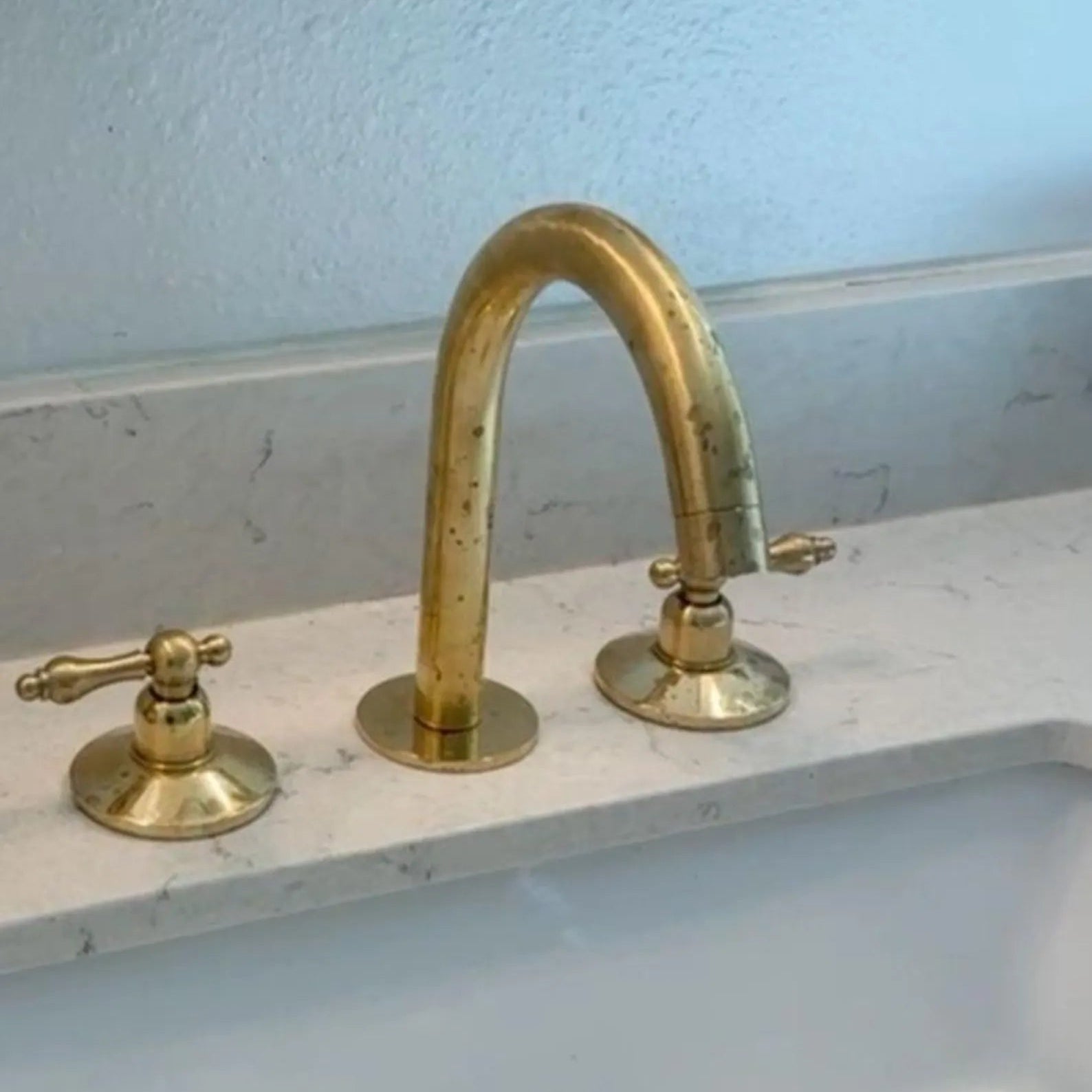 A close-up of a bathroom sink with a curved gold faucet, two matching gold handles, a white sink basin, and part of a mirror above.