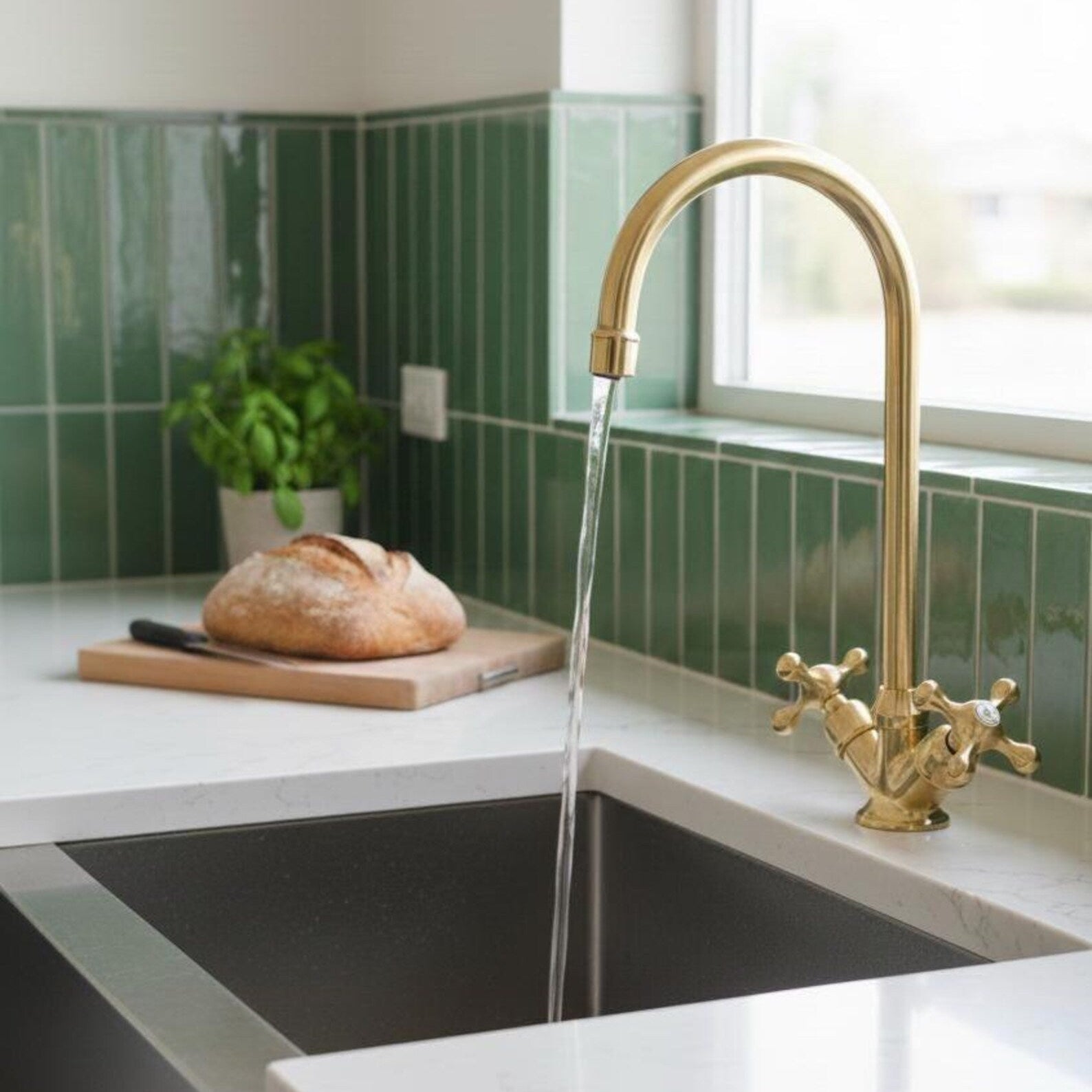 A modern kitchen scene featuring a tall, arched matte gold faucet with water running from the spout into a dark sink, set against a green tiled backsplash and white countertop with a loaf of bread on a cutting board.