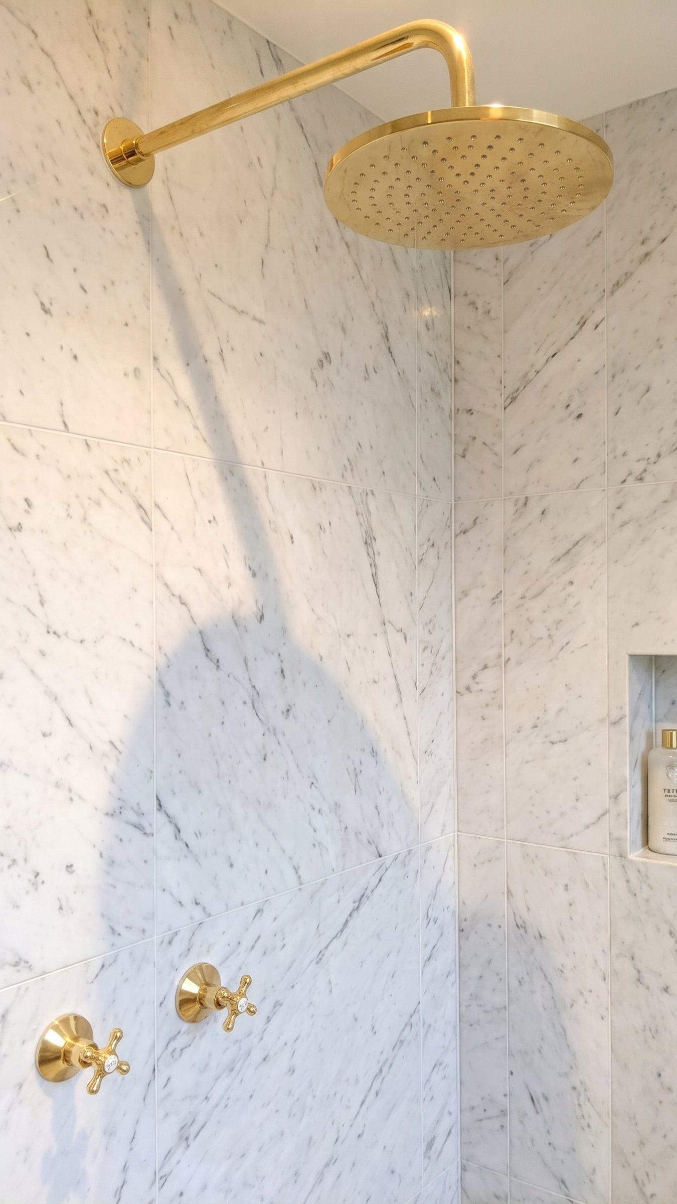 A modern bathroom corner featuring a large round gold rain shower head mounted on a curved gold arm, set against white and gray marble tiled walls with matching gold controls and a built-in niche shelf.