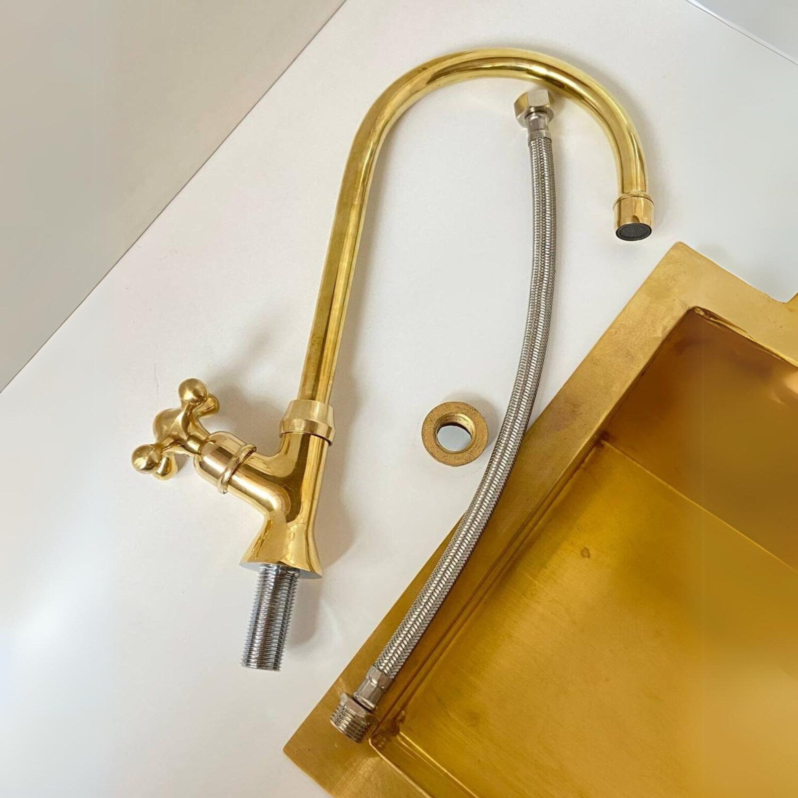 A brass faucet assembly with a curved gooseneck spout, cross-handle valve, exposed threaded pipe, and a braided metal supply hose, shown on a white surface next to a matching brass sink basin.