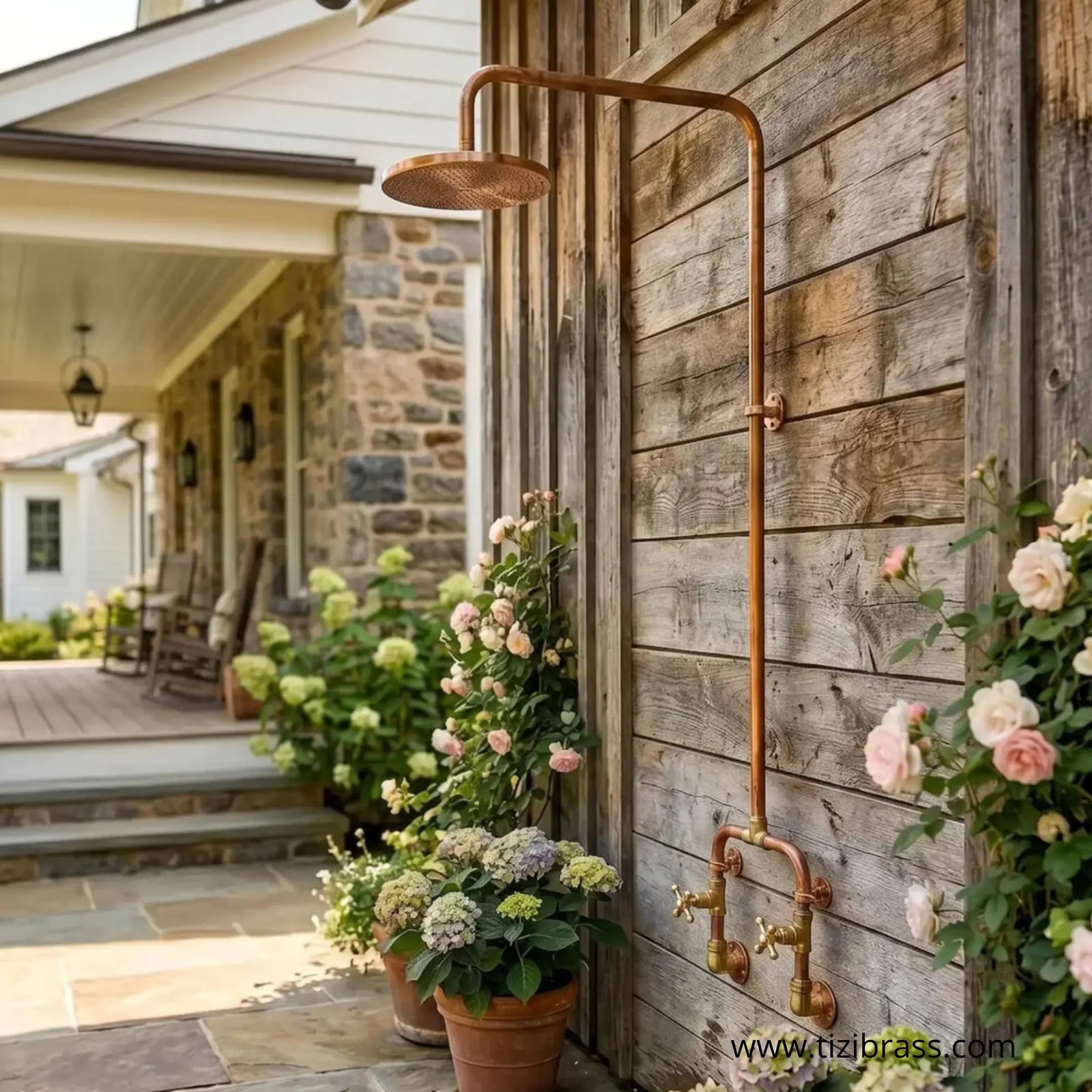 Unlacquered brass outdoor shower mounted on a wooden wall next to potted flowers and a stone patio.