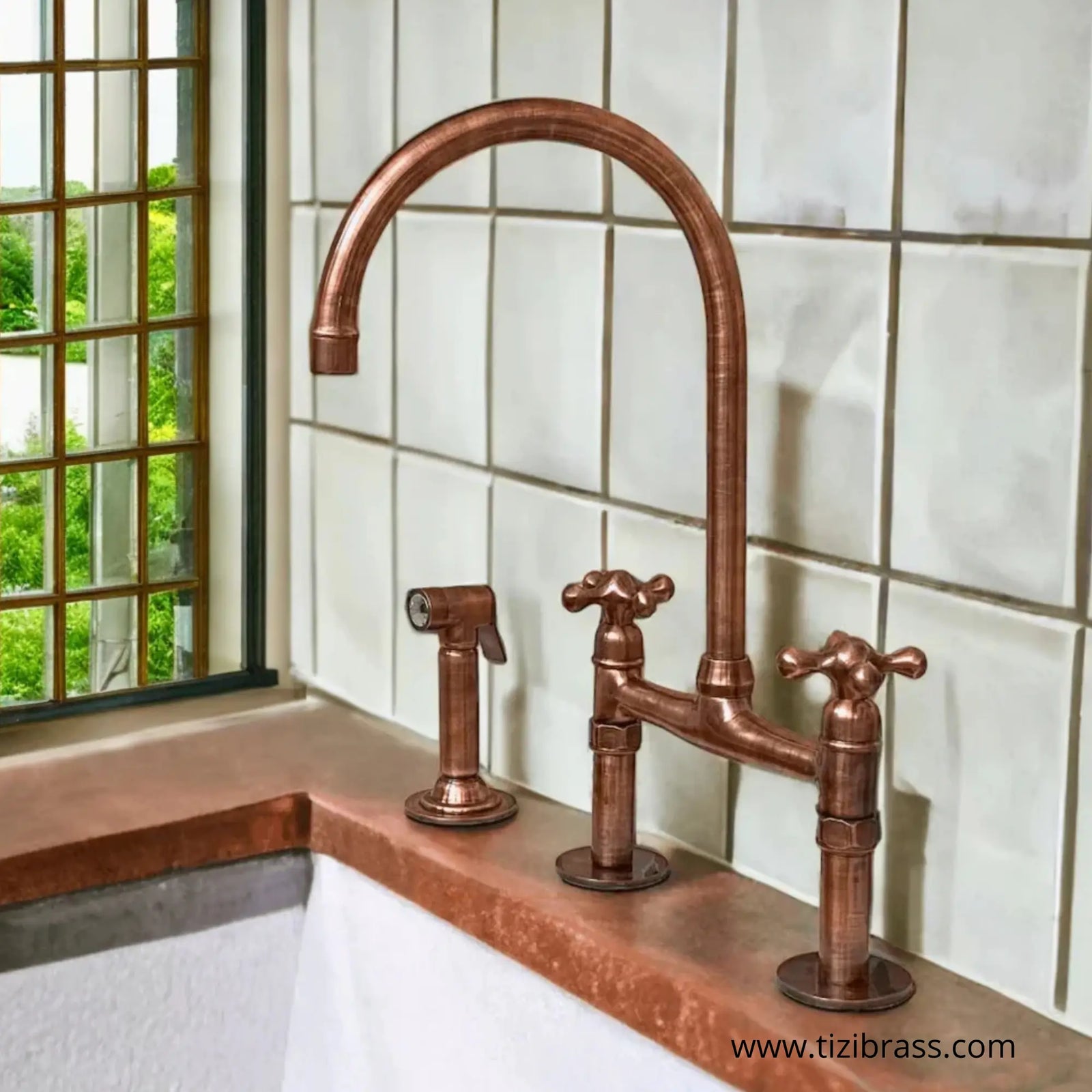 Antique unlacquered copper bridge kitchen faucet with cross handles, matching side sprayer, and high-arc swivel spout on a white tiled backsplash.
