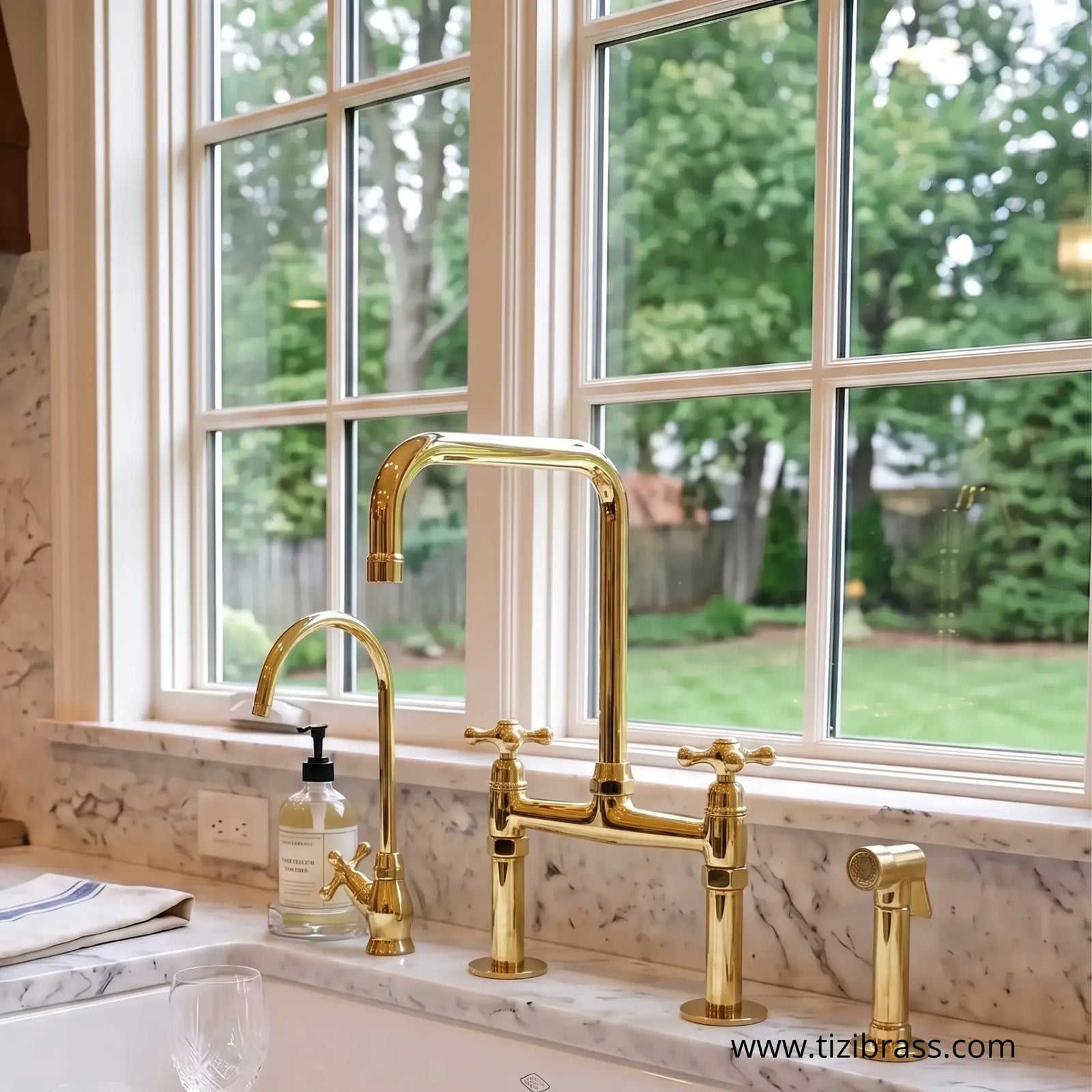 Unlacquered polished brass bridge kitchen faucet with cross handles, gooseneck spout, filtered water tap, and side sprayer on a white marble countertop beneath a window.
