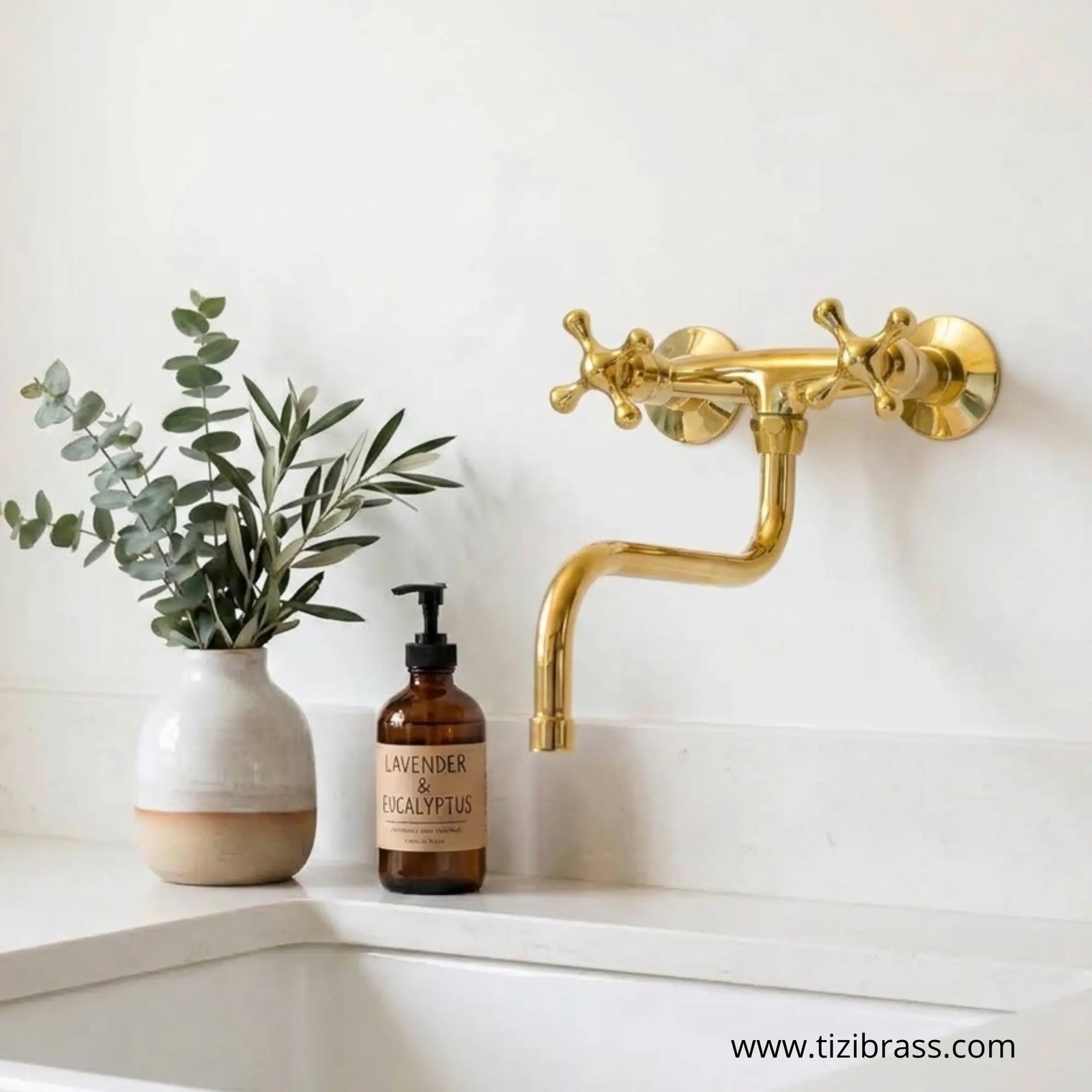 Polished brass wall-mounted faucet with cross handles installed on a white wall above a sink, featured next to greenery and an amber soap bottle.