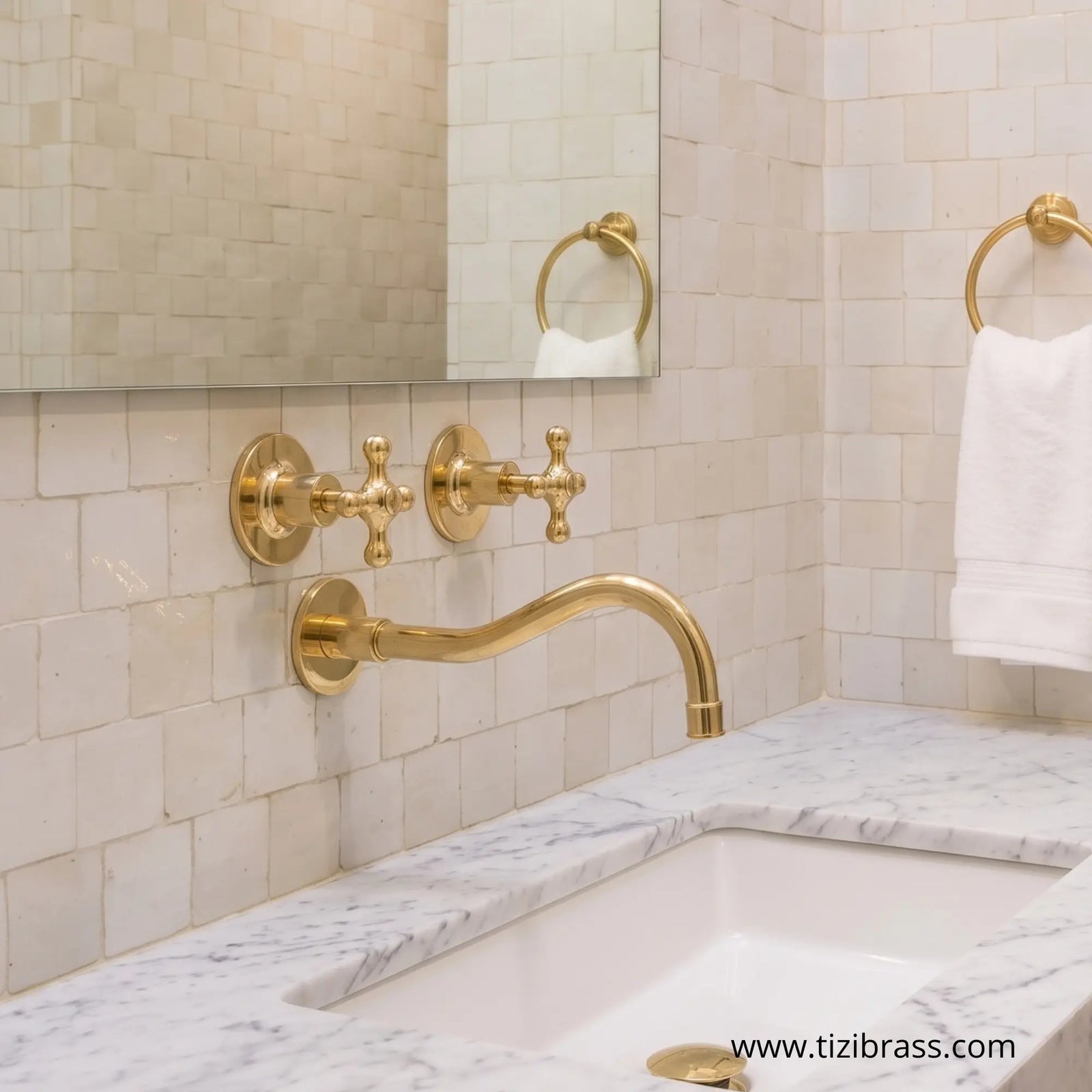 Unlacquered wall-mounted brass faucet with cross handles over a marble sink and zellige beige tile backdrop.